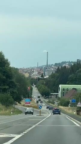 The video shows a car's point of view traveling on a paved road that curves to the left. The road has multiple lanes and white dashed lines dividing them. Several cars are visible on the road ahead, moving in the same direction. On the left side of the road, there are trees and a Swiss flag is planted on a pole. There is also a billboard with pink and blue graphics. On the right side of the road, there are more trees, some buildings, and a fence. In the distance, a city with buildings and a tall industrial chimney with smoke coming out of it is visible on a hillside. The sky is overcast. The overall scene is a view of a highway leading towards a town with industrial elements.