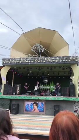 The video shows a band performing on an outdoor stage. The stage has a large, horn-shaped backdrop decorated with musical notes. There are four band members on stage: one playing a bass guitar, one singing and playing a guitar, one playing drums, and another playing a guitar. The band is illuminated by stage lights. In front of the stage, there is a large display featuring a portrait of a woman. Spectators are visible in the foreground, with the backs of their heads facing the camera. The sky is overcast.