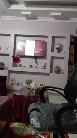 The video is shot indoors in what appears to be a living room. The walls are painted a light purple color and feature recessed shelves displaying decorative items. A flat-screen television is mounted on the wall between the shelves, showing various screens throughout the video. Below the TV is a table covered with a red cloth. A black office chair is positioned in the foreground, partially obscuring the view. A checkered shirt is draped over the back of the chair.  The room also contains a red couch. The ceiling has white rectangular insets. An Arabic script is hanging from the wall above the doorway.