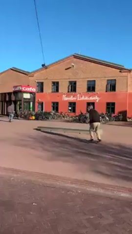 The video shows a person skateboarding on a brick-paved plaza under a clear blue sky. The skateboarder, wearing dark clothing and light-colored pants, rides towards a concrete ledge. They perform a trick, appearing to attempt to grind on the ledge. Several other people are present in the background, some walking, others standing or sitting near a building with the words "NORREBROHALLEN" on it. There's also a cafe visible, along with numerous bicycles parked nearby. The camera follows the skateboarder, capturing the trick and the surrounding environment.