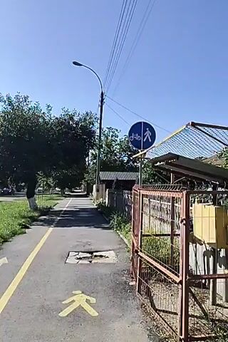 The video shows a paved path with a yellow line dividing it, suggesting a bike lane and pedestrian area. A sign at the beginning of the path indicates it is for both cyclists and pedestrians. There is a large pothole in the path, with broken pavement around it. A manhole cover is visible within the pothole. A fence runs along the right side of the path, and trees and greenery are visible on the left. The sky is clear and blue. The camera is moving forward along the path.