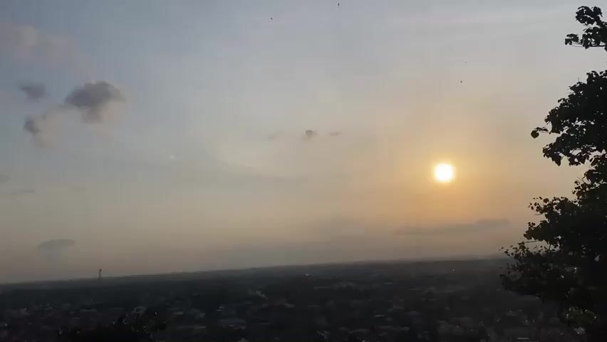 The video shows a panoramic view of a cityscape from an elevated viewpoint, likely a hill. The sun is setting, casting a warm, golden glow across the sky, which is partly cloudy with some fluffy white clouds and some lighter grey ones. The sun is a bright, orb-like disc in the upper right quadrant of the frame. Below the horizon, a dense urban area is visible, with many buildings and homes stretching out as far as the eye can see. The colors of the city are muted by the fading light, appearing as a dark tapestry of rooftops. The foreground features the dark silhouettes of trees and foliage, indicating that the viewpoint is situated in a natural, possibly hilly, area overlooking the city. The overall atmosphere is serene and peaceful, capturing the beauty of a sunset over a populated area. There are no people or distinct actions visible, only the static environmental scene.