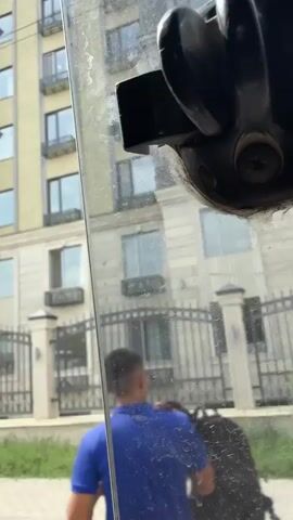 The video shows a close-up of a black handle on a glass surface, possibly a car window. In the background, a modern apartment building with several stories and windows is visible, along with a black metal fence and a light-colored car parked in front of the building. The camera appears to be inside a vehicle looking out.