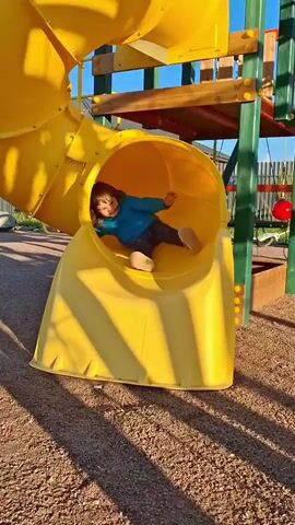 A young child wearing a blue hoodie and patterned leggings is playing on a yellow playground slide. The child slides down the tunnel and then walks away from the bottom of the slide. The playground appears to be made of wood and metal, with a gravel surface. In the background, a house and trees are visible. The sun is casting long shadows across the ground, indicating it is likely late afternoon.