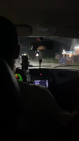 The video shows the interior of a car at night. The camera is positioned behind the driver, facing forward. The dashboard and windshield are visible. A person's head and shoulder are in the foreground, obscuring part of the view. The driver's hands are on the steering wheel. There is a phone mount with a phone on the dashboard. A rearview mirror is also visible. Outside the car, lights and street signs are faintly visible in the darkness. The video appears to be a recording from inside a moving vehicle.