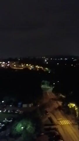This is a low-light video taken at night from a high vantage point, possibly a building. It shows a landscape with scattered lights, suggesting a residential or urban area. In the foreground, a crosswalk is illuminated by a streetlight. The overall scene is dark with the only visible light sources being artificial lights from buildings and streetlights. The sky is overcast.