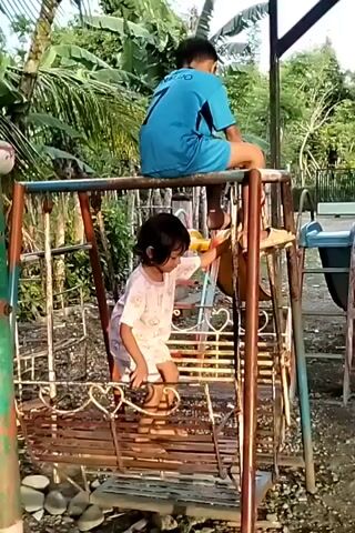 The video shows two children on and around a swing set in an outdoor environment. A young girl with short dark hair, wearing a light-colored shirt and shorts, is inside the swing. She is looking to her right and appears to be reaching out with her right hand. A boy wearing a blue shirt and blue shorts is sitting on top of the metal swing set structure. He is facing away from the camera. The swing set itself appears to be made of metal and shows signs of rust and wear. Behind the swing set, there is lush green vegetation, including palm trees. To the right of the swing set, there is another piece of playground equipment and a black fence. The ground around the swing set is covered in gravel. The video is taken during the day, and the lighting suggests it might be late afternoon.