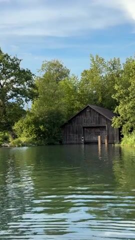 The video shows a view of a lake with a wooden boathouse on the right side, surrounded by trees. The water is calm and reflects the sky and trees. The boathouse is dark and appears weathered, with a large opening for boats. There is a large, leafy tree on the left side of the frame, near the shore. The sky is blue with some white clouds. The overall scene depicts a serene natural environment.