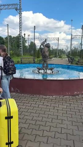 A bright daytime video shows a group of people at what appears to be a railway station or an area adjacent to train tracks in Malaya Vishera, Russia. The sky is blue with scattered white clouds. The scene includes a large circular fountain with a red base and blue water, featuring a statue of a cherub-like figure spraying water. Overhead railway electrical lines and structures are visible above the tracks in the background.

Several individuals, including adults and children, are walking along a paved path, pulling various suitcases. One person in the foreground pulls a bright yellow suitcase past the fountain in the opening shot. Later, a wider shot shows multiple people, some with backpacks (including a black one), pulling different colored suitcases, such as another yellow one and a maroon one. Benches are positioned along the paved area, and greenery lines the background.

The video concludes with a focus on a young girl with red hair, who is smiling happily as she pulls a small yellow suitcase. She wears a black shoulder bag with "HELLO KITTY" written on it. The overall atmosphere suggests people traveling or arriving at a destination.
