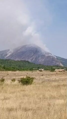 The video shows a mountain with smoke rising from its summit, indicating a fire. The mountain is partly covered in vegetation, with some areas appearing burnt or scorched. In the foreground, there is a field of dry, yellow grass with a few bushes. The sky is partly cloudy. The video pans across the landscape, revealing more of the mountain and the surrounding vegetation. There are no people visible in the video.