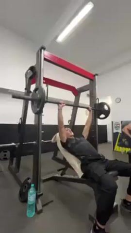 A man is performing a bench press exercise in a gym. He is lying on a weight bench placed inside a Smith machine, gripping the barbell. He lowers the barbell towards his chest and then pushes it back up. He’s wearing black workout clothing. There is a water bottle and weights on the floor. The gym has white walls and a black and red Smith machine. There is another man visible in the background.