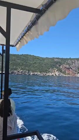 The video shows a view from a boat moving on the sea. The water is blue and calm with small waves. The boat has a white canopy overhead. In the distance, there is a coastline with green hills and mountains. The sky is clear and blue. There are no people clearly visible in the frame. The boat is moving forward, creating a wake in the water. The camera pans slowly to the right, revealing more of the coastline and the sky. The lighting suggests it is daytime, possibly late morning or early afternoon.