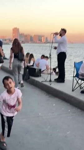 A vertical video shows an outdoor scene with a body of water on the right and a walkway on the left.  In the foreground, a young boy wearing a pink t-shirt and black shorts dances with his arms outstretched, looking at the camera. He has dark hair and is wearing dark sandals.  Further back along the walkway, a man in a white shirt and black pants stands and sings into a microphone on a stand. He is holding a phone or small tablet in his left hand.  Behind him, several people are sitting on the ground facing the water, some in white clothing. A black speaker sits on the ground near the singer. In the background, a city skyline with tall buildings is visible across the water, bathed in the warm light of sunset. More people are walking along the distant walkway. The sky is a soft orange and pink.