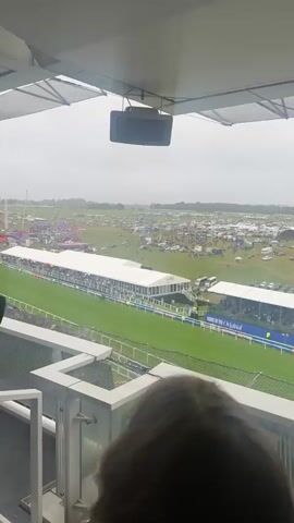 A high-angle shot from an elevated position overlooks a horse race on a cloudy day. In the foreground, the back of a person's head with brown hair is visible behind a metal railing. To their left, a person wearing a dark suit jacket and white shirt enthusiastically waves a dark hat, appearing to cheer. On the green race track below, several horses with jockeys are racing from left to right. A long white tent structure filled with spectators runs parallel to the track. Beyond the track, a large open field contains numerous parked vehicles and smaller tents with many people. A black speaker is mounted above the camera's view. The audio includes cheering and race commentary.