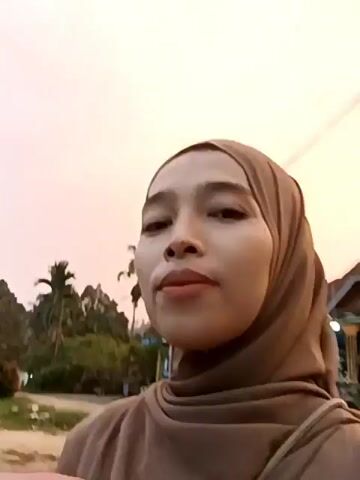 A woman wearing a hijab is holding a child who is wearing a pink hoodie. The woman is smiling and looking at the camera. The child is looking away from the camera. The background is blurred and appears to be outdoors with trees and a building. The lighting suggests it is either sunrise or sunset.