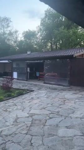 The video presents a panoramic view of a residential property, likely a backyard or rural estate, in Rainovtsi, Bulgaria. The scene unfolds under a soft, diffused light, suggesting either early morning or late evening, with a sky showing hints of pink and blue through light clouds.

The camera pans from left to right, starting with a stone-paved patio area. To the left, a building with a dark roof is visible, featuring a covered outdoor seating area or gazebo with curtains. Adjacent to the main building, there appears to be an open-sided structure, possibly an outdoor kitchen or garage, with transparent screens. The stone patio is made of irregularly shaped flagstones.

As the camera continues to pan, it reveals a well-maintained grassy lawn bordered by garden beds with various plants and potted flowers. In the distance on the lawn, a blue rectangular object and a trampoline with a safety net are visible. Further along, a wooden fence and a decorative wooden gate define the property's boundary. Beyond the fence, a utility pole with power lines stands tall, and a white car is briefly seen on what appears to be a road. The background consists of lush green trees and distant hills or mountains under the soft twilight sky, creating a peaceful and serene ambiance. No people or visible text are present in the video.