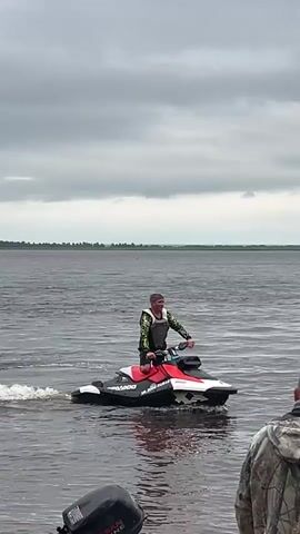 A person rides a red and white jet ski on a body of water. The person is wearing a life jacket over a camouflage shirt and dark shorts. The water is dark and choppy. In the foreground, the back of another person wearing a camouflage jacket is visible, partially obscuring a black outboard motor attached to a boat. The background shows a distant shoreline with trees under a cloudy sky. The jet ski is kicking up a wake. The person on the jet ski is looking towards the camera and appears to be talking or gesturing.