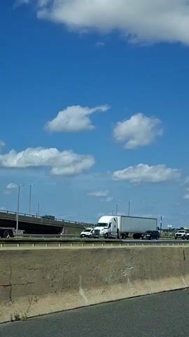 The video shows a view from a moving vehicle on a highway. The sky is blue with scattered white clouds. On the highway, there are several vehicles including semi-trailer trucks, cars, and SUVs. The vehicles are moving in the same direction. In the background, there are some buildings and a bridge-like structure. An airplane is visible in the sky in the distance. The concrete barrier of the highway is in the foreground. The video appears to have been shot during the daytime.