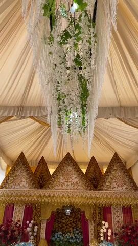 The video shows a decorated wedding stage. The stage is adorned with traditional triangular roofs in gold and white, with intricate patterns. Red curtains are draped on the sides of the stage. There are floral arrangements on both sides of the stage, with red and blue flowers. Hanging from the ceiling is a large floral arrangement of white flowers and greenery, with a beaded chandelier-like structure. The entire setup is covered by a cream-colored tent.