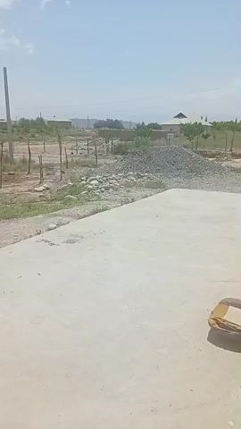 This video captures an outdoor scene on a bright, sunny day with a clear blue sky dotted with a few white clouds. The foreground is dominated by a large, light-colored concrete surface, possibly a patio or foundation. To the right of the concrete, a tan object, possibly a bag or cushion, rests near the edge.

The middle ground reveals a dusty, arid landscape with scattered patches of green vegetation and some dry grass. Several wooden stakes are driven into the ground, suggesting a boundary or construction area. A pile of grey gravel sits prominently in the middle right of the frame. Beyond the stakes and gravel, more trees and bushes are visible, interspersed with what appear to be simple, one-story buildings with light-colored roofs. In the distance, a line of hills or mountains can be seen under the hazy sky. A tall, grey utility pole stands prominently on the left side of the frame, and another similar pole is visible further back. The overall impression is of a rural or semi-developed area, possibly undergoing some form of construction or landscaping. The bright sunlight and clear sky suggest it is midday. There is no visible text in the video.