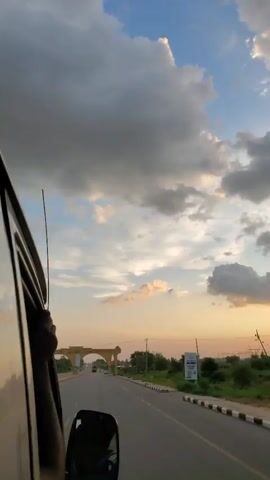 A first-person perspective video captures a view from inside a vehicle, looking out the passenger side window. The sky is filled with dramatic clouds, with a clear blue sky visible above. The vehicle is traveling on a paved road that leads towards a large, arched gateway. The gateway has text written on it, which appears to say "JUJULDO TARABA STATE CAPITAL". The gateway is illuminated by the setting sun, casting a warm glow. Trees and power lines line the sides of the road. The video is in portrait orientation and lasts for five seconds.