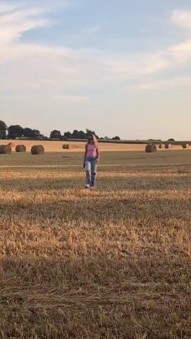 A young woman with blonde hair, wearing a pink tank top and ripped blue jeans, walks across a harvested field. She is in the center of the frame, facing the camera. The field is covered in dry, cut straw. In the background, several large, round hay bales are scattered across the field. The sky above is a pale blue with wispy clouds, and the sun appears to be low on the horizon, casting a warm light on the scene. Trees line the far edge of the field. The overall atmosphere is serene and rural.