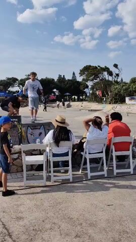 The video shows a skateboarding event outdoors. In the foreground, a young boy wearing a blue baseball cap and a graphic t-shirt is walking left to right. Behind him, several people are seated in white chairs, some wearing hats and casual clothing. A stone wall separates the seated spectators from the skateboarding area. In the background, a skate park is visible with ramps and concrete surfaces. Several skateboarders are present. One skateboarder in a white t-shirt and shorts is holding a skateboard and looking to the left. Another skateboarder in a black t-shirt is walking towards the center of the frame. Further in the background, other individuals are skateboarding or standing around the park. The sky is blue with scattered clouds, and trees and buildings are visible in the distance. The overall scene is of a sunny day at an outdoor recreational event.