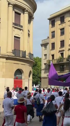 A group of people in white shirts and red or blue accents are walking down a cobblestone street between tall buildings. Most of them are facing away from the camera and moving towards the right of the frame. Several individuals are carrying a large purple flag with an emblem on it. One person in the foreground is carrying a blue bucket on their shoulder. The buildings appear to be old European architecture with balconies and windows. The sky is visible at the top of the frame and appears partly cloudy. The overall atmosphere seems to be festive, possibly a parade or procession.
