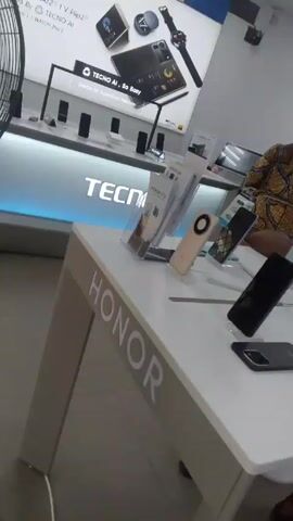 The video shows the interior of a mobile phone retail store. There are multiple display tables showcasing different brands, including Tecno, Honor, Oppo, and Infinix. Sales representatives and customers are present in the store. The lighting is bright, and promotional posters for various phone models are visible on the walls. The camera moves from one display table to another.