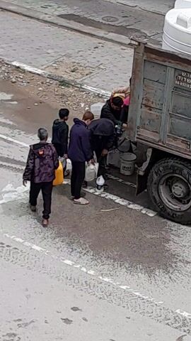 Several people are gathered around the back of a large, old, and weathered truck. They appear to be filling containers with liquid from a spigot or opening on the truck's rear. The containers vary in size and type, including jugs, bottles, and buckets. The ground around the truck is wet, suggesting spillage. The surrounding environment looks like an urban street, with pavement and some visible buildings in the background. The scene gives the impression of a communal water or liquid distribution point.