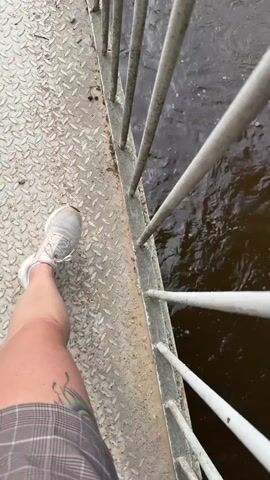 A first-person perspective video shows someone standing on a bridge over a body of water. The person's left leg and foot, clad in a white sneaker and plaid shorts, are visible on the left side of the frame. The bridge has a diamond-plate metal walkway and a metal railing that runs along the edge. The water appears murky and is flowing. Green foliage hangs over the edge of the bridge on the right side of the frame. The camera moves slightly, giving a downward-facing view of the scene.
