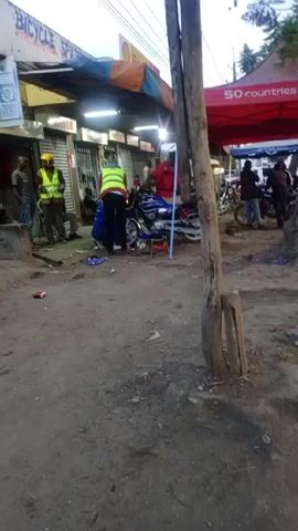 The video shows a street scene. On the left side, there's a building with a sign that says "BICYCLE SPARES." People are gathered around a blue motorcycle, seemingly working on it. One person wears a yellow vest, while another wears a red jacket. The ground is dirt and there's scattered debris.

On the right side, a red tent with "50 countries hot sale" written on it is visible. People are standing around under the tent. A large tree stands next to the tent. A street runs along the right side of the frame. The weather appears overcast.