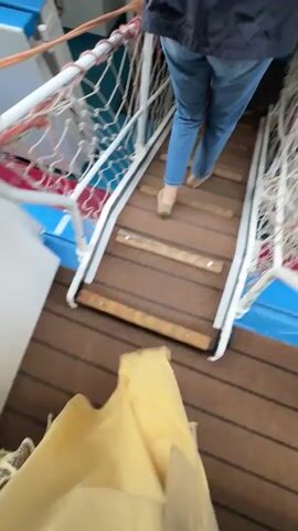 This video shows a person walking down a set of wooden stairs with rope railings on the sides. The stairs appear to be on a boat or ferry, with blue and white walls visible. Another person in blue jeans and a dark jacket is walking up the stairs ahead of the camera person. The camera person is wearing a tan-colored garment that covers their legs. The stairs are wet in places and have some visible scuff marks. The overall environment appears to be functional and utilitarian.