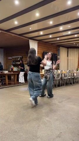 This video depicts two young women joyfully dancing in a spacious, well-lit indoor venue, likely a common hall or dining area, located in Yus'ki, Russia. The woman on the left has long, dark and light two-toned hair, and is dressed in a black t-shirt, baggy light blue jeans, and white sneakers. Her companion, on the right, has long dark hair and wears a white tank top, a plaid shirt tied around her waist, baggy light blue jeans, and dark sandals. Both women are actively dancing, smiling, and appear to be having a great time, moving energetically to the music playing in the background.

The room has a rustic yet well-appointed feel, featuring wooden paneled walls and a white ceiling with exposed wooden beams and modern recessed lighting. On the right side of the frame, multiple tables are set with chairs, suggesting a dining or social gathering. On the left, a long wooden table laden with various drinks and snacks is visible, with another woman casually dressed in white pants and a dark top standing behind it. White curtains are draped along the walls, and string lights are visible in the background, contributing to a festive atmosphere. The floor is carpeted. The overall scene conveys a cheerful and relaxed social event, possibly in the evening given the reliance on artificial lighting. No discernible text is visible within the frame.