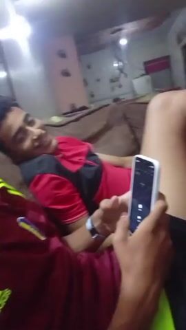 Two people are sitting on a couch in a living room. The person holding the phone is wearing a maroon Adidas shirt with yellow stripes, and is looking down at the phone. The other person, leaning back on the couch, is wearing a red shirt with a black stripe. The living room has light pink walls and a dark brown couch. A light fixture is hanging from the ceiling. The person holding the phone has a smartphone in their hands, possibly an iPhone, and appears to be using it. At one point, the phone’s camera lens is obscured by the person’s hand.
