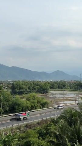 A highway stretches across the middle of the frame, carrying a few vehicles including a green truck with a red tarp and a white van. To the left of the highway is a lush green area with trees and some buildings, and to the right are flooded rice fields and more greenery. In the distance, a range of mountains forms the horizon under a cloudy sky. Palm fronds are visible in the foreground.