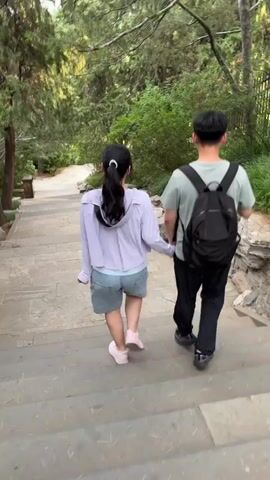 A young couple walks hand-in-hand up a stone staircase in a park. The woman, on the left, wears a light purple zip-up hoodie, a light blue shirt, and denim shorts. Her dark hair is pulled back in a ponytail. The man, on the right, wears a light green t-shirt, black pants, and a black backpack. They are surrounded by lush green trees and foliage, with large rock formations visible on the right side of the path. The camera is positioned behind the couple, capturing their ascent.