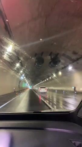 The video shows the interior of a car driving through a tunnel. The tunnel is illuminated by lights along the ceiling and walls. The road surface is wet, indicating that it is raining or has recently rained. Several cars are visible in front of the vehicle, including a white sedan. The perspective is from the driver's seat, looking forward through the windshield. The windshield itself appears to be dirty or streaked, with some reflections and smudges visible. The overall impression is of a dark, enclosed space with artificial lighting.