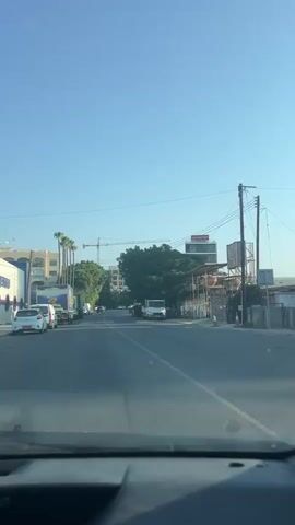 The video shows a view from inside a vehicle driving down a street. The sky is clear and blue. On the left side of the road, there are several parked trucks, some with large colorful advertisements on their sides, and some smaller cars. Palm trees and buildings are visible behind the trucks. On the right side of the road, there are trees, a white truck, and a covered structure with some equipment or goods underneath. There is also a utility pole with wires running across. Further down the street, there are more vehicles, including cars and trucks, and some buildings in the distance. A construction crane is visible in the background. The road has a white line marking the center. The video is shaky, indicating movement.