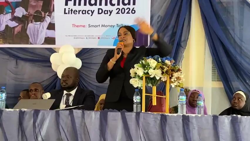 The video captures an indoor event, likely a seminar or conference, featuring a speaker and seated panelists. A woman, dressed in a black suit with a red inner top, is the central figure, actively speaking into a microphone and gesturing with her right hand. She stands behind a long table covered with a blue and white patterned tablecloth.

On the table, in front of the speaker, are two bottles of water. To her right, a floral arrangement with white and blue flowers is displayed on a gold-colored stand. To her left, a cluster of white balloons is visible behind the table.

Seated behind the table are several individuals. To the speaker's left, a man in a dark suit and tie is attentive, with an open laptop in front of him. Another man is partially visible next to him. To the speaker's right, three individuals, likely women, are seated, wearing headscarves (hijabs), also listening intently.

In the background, a large banner is displayed, featuring the text "Financial Literacy Day 2026" and the "Theme: Smart Money Talks." Below the theme, smaller text and social media handles like "@NDICNigeria" are visible. Blue drapes flank the banner, completing the formal setting. The speaker appears animated and engaged with her audience.