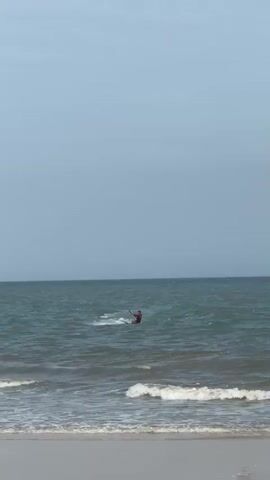 The video captures a wide shot of a serene ocean with a hazy sky. In the foreground, gentle waves lap at a sandy shore. Further out, a kiteboarder navigates the water, their board creating a splash as they move. A person is visible in the water to the left of the kiteboarder. The kiteboarder appears to be performing a jump or maneuver, with their board briefly lifted out of the water. The scene is calm and suggests a day suitable for water sports.