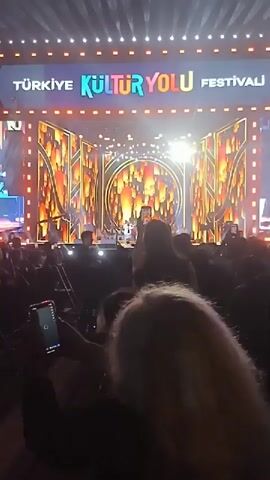 The video shows a performance taking place at the Türkiye Kültür Yolu Festivali. An audience is seated in front of a stage, recording the performance with their phones. The stage is decorated with lights and patterns.