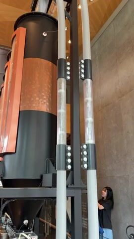 The video shows a tall, black and copper industrial machine with two clear tubes running vertically beside it. Inside the tubes, small, round objects are visible, some clear and some appearing to be small, light-colored spheres. There are lights or sensors attached to the tubes at intervals. On the right side of the frame, a young woman with dark hair is standing and looking at the machine. She is wearing a dark t-shirt and a blue jacket. In the center of the frame, another person, possibly a man with lighter hair, is partially visible between the tubes. In the background, a rough-textured wall and a wooden ceiling are visible, suggesting an indoor environment, likely a public space like a coffee roastery or a themed attraction. The woman is gesturing towards the machine, and her mouth is open, as if expressing surprise or excitement.