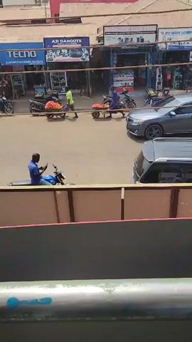 The video shows a street scene in what appears to be a commercial area, with various shops visible along the side of the road. Several people are visible. One man sits on a motorcycle looking at his phone. Another man walks across the street, pushing a cart filled with red items that may be tomatoes. Motorbikes are parked on the side of the road. Cars are parked and moving on the street. The video appears to be taken from a building overlooking the street.