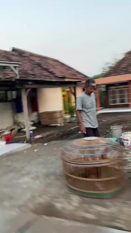 The media file depicts an outdoor scene in Sumorame, East Java, likely during the late afternoon or early evening given the soft, diffused light. A person, dressed in a grey t-shirt and dark pants, is visible in the mid-ground, appearing to be walking or standing near a house.

The setting is characterized by a single-story building with a tiled roof and a light-colored facade. A bright yellow door with blue accents is a prominent feature of the house. Various objects are scattered around the foreground and mid-ground, including several bird cages of different sizes and materials, some appearing to be made of bamboo or wire. Other items, like buckets and what might be discarded materials, are also present.

The overall impression is of a residential area, possibly a street or yard, with a somewhat cluttered but lived-in feel. The motion blur suggests the footage was taken from a moving vehicle. There is no discernible text visible in the frame. The weather appears to be calm, with no strong indicators of wind or precipitation. The scene evokes a sense of everyday life in a rural or semi-urban Indonesian setting.