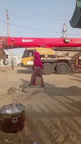 A woman wearing a pink hijab and carrying a shopping bag walks in front of a large yellow and red SANY crane. The crane is parked on a sandy street. A man walks in the background. In the foreground, a weathered, metal container with a lid sits on a gravelly surface. Power lines and a cell tower are visible in the distance. The sky is clear and blue.