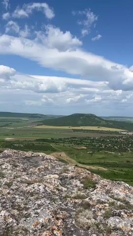 The video shows a landscape viewed from a rocky outcrop.  Below is a green, cultivated valley with a village.  In the distance, the land stretches to the horizon under a partly cloudy, blue sky. The camera pans slowly.