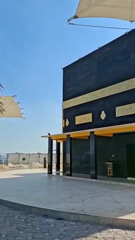 The video opens with a view of a black building with gold accents, resembling the Kaaba in Mecca. A car is parked in front of the building. A palm tree is visible to the left of the building. The sky is clear and blue. The camera pans to the right, showing more of the building and a large white tent-like structure overhead. The video then shows a close-up of the building, highlighting its architectural details. The camera continues to pan, revealing more of the surrounding area, including a minaret-like structure and other tent-like canopies. A white van is parked in the distance. The video ends with a shot of the sky and the canopies.