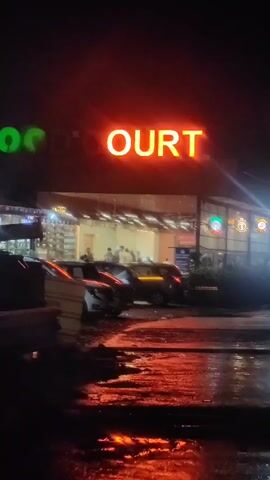 The video shows a street scene at night, possibly after rain, with reflections on the wet ground. A large red neon sign that reads "COURT" is prominently displayed above a building with people inside. Several cars are parked in front of the building. The overall atmosphere is dimly lit and appears to be urban.