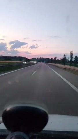 The video is taken from the perspective of a driver traveling on a highway at dusk. The highway has multiple lanes and is bordered by fields and trees. The sky is a gradient of pale pink and blue, with a few wispy clouds. There are other vehicles on the highway, their headlights visible in the distance. In the foreground, a portion of the car's dashboard is visible, including a black electronic device, possibly a GPS or dashcam. The overall mood is peaceful and serene, evoking a sense of a journey at the end of the day.